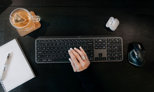 a person typing on a keyboard with headphones, a mug, and pen and paper 